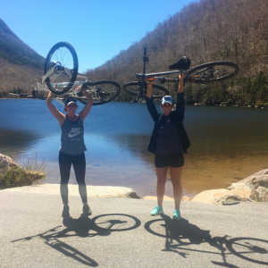 Kate Martin and her friend holding their mountain bikes in font of a lake.