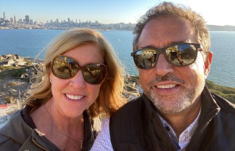 Photo of a man, David Almeda, and his wife, Erin wearing sunglasses and water and city skyline standing in front of a view of the 