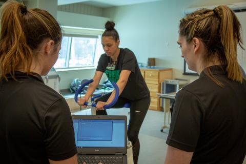 Two female students looking at a laptop screen face a female student on a Stairmaster.