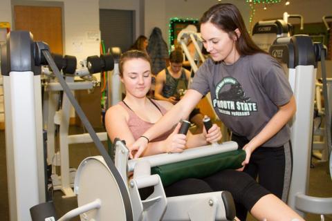 A female campus recreation student employee helps a female student use a weight machine in the Plymouth State University weight room.