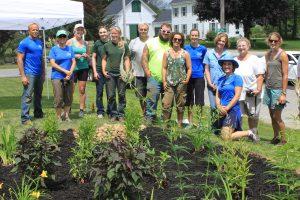 A group of faculty & staff stand together in front of a garden with tools in hand on Plymouth State University's campus