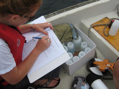 A faculty member sits on a canoe in water with a box of water samples next to her while she documents findings on a piece of paper in a 3-ring binder.