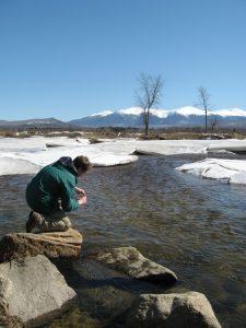 A student kneels on a rock at a river with snow capped mountains in the background.