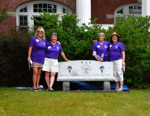 Four women wearing matching purple shirts stand in front of a granite bench